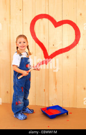 smiling little girl with a brush in hand standing in front of a wooden wall on which she has painted a heart with red paint Stock Photo