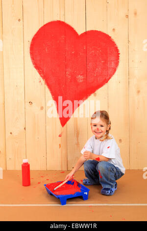 laughing little girl squatting in front of a wooden wall on which she has painted a heart with red paint Stock Photo