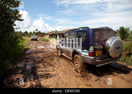 cross-country vehicles driving through a mud pit on a soil road through bush landscape passing humble huts, Burundi, Cankuzo, National Parc de la Ruvubu, Cankuzo Stock Photo