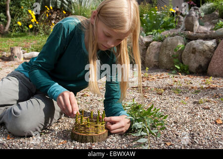girl building an Easter basket from a tree disc, willow twigs, moss, daisies and coloured eggs; 3. step: weaving flexible willow twigs into the solide vertical ones, Germany Stock Photo