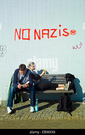 young couple on a bench in front of a concrete wall with the writing 'NO NAZIS' sprayed upon Stock Photo