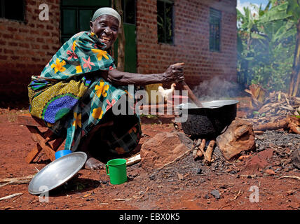 happy old woman is cooking rice on an open fire place in front of her home, Uganda, Jinja Stock Photo
