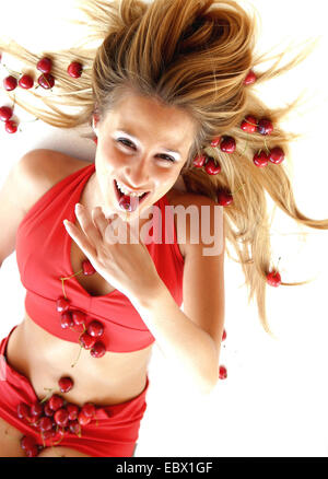 young woman lying on the ground with flowing hair covered with cherries and eating one of them with a smile Stock Photo