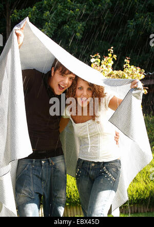 young couple standing in the garden in a rain shower laughing while holding a tablecloth over their heads Stock Photo