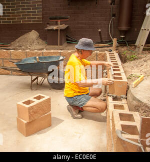 Man wearing bright yellow shirt, shorts, and hat laying coloured concrete blocks to create wall on concrete slab Stock Photo