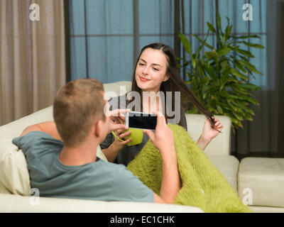 young man sitting on sofa taking a photo of his girlfriend at home Stock Photo