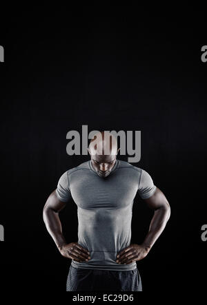 Portrait of muscular young man in sportswear standing with his hands on hips looking down against black background. Stock Photo