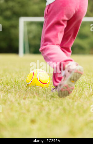 Girl in pink pants playing soccer, kicking a yellow football in front of goal. Stock Photo