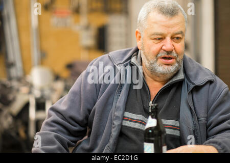 An older caucasian man between 60 and 70 years sitting in his shop with beer bottle talking to somebody. Stock Photo