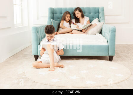 Siblings spending free time at home: two girls reading book on couch and boy using tablet while sitting on floor Stock Photo