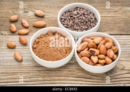 cacao beans, nibs and powder in white ceramic bowls against grained wood Stock Photo