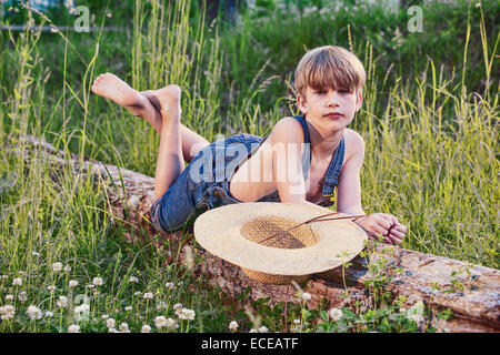 Young boy wearing blue overalls laying on fallen tree Stock Photo