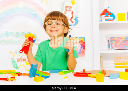 Cute little boy wearing glasses holding plastic hammer wrench and screwdriver, sitting in the kindergarten class with other tool Stock Photo