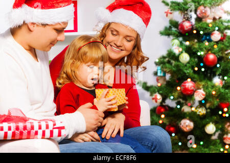 Family with mom dad and little three years old daughter sitting near Christmas tree and opening New year presents surprised litt Stock Photo