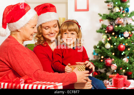 Family with mom granny and little three years old daughter sitting near Christmas tree and opening New year presents with smile Stock Photo