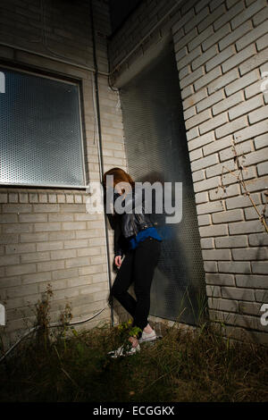 Victim of abuse: A sad depressed afraid scared solo redhead ginger haired young woman teenager girl alone by herself  leaning on a wall outside a derelict abandoned block of flats UK