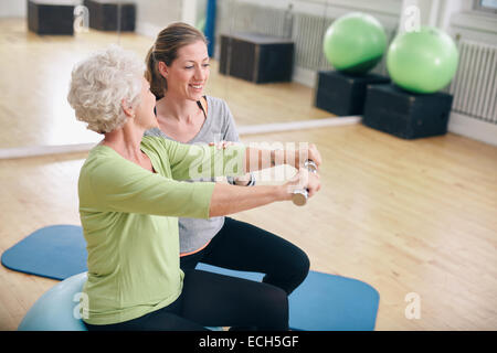 Senior woman exercising with weights in the gym assisted by a young female trainer. Old woman lifting dumbbells with help. Stock Photo