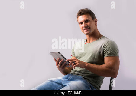 Muscular young man sitting on chair reading from ebook device, smiling at camera Stock Photo