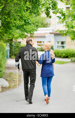 Full length rear view of female caretaker walking with disabled senior man on street Stock Photo