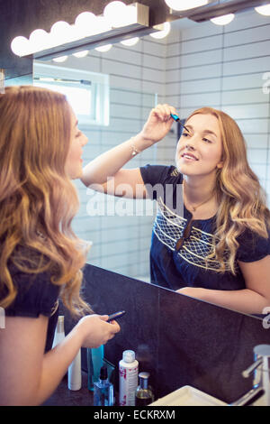 Happy young woman applying mascara in front of bathroom mirror Stock Photo