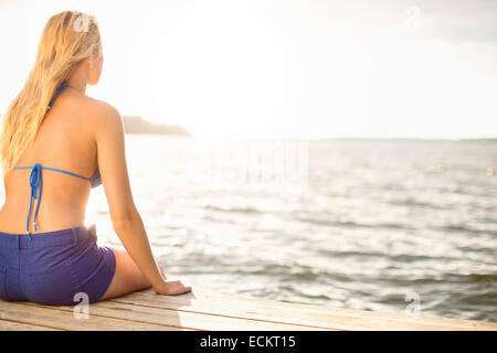 Rear view of woman in bikini top sitting on boardwalk by lake against sky Stock Photo