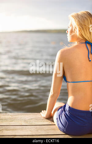 Rear view of young woman in bikini top sitting on boardwalk by lake Stock Photo