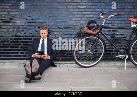 Full length of businessman using digital tablet while sitting on sidewalk by bicycle Stock Photo