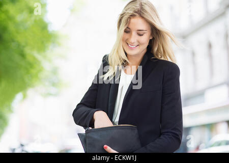 Smiling young woman searching something in her clutch bag outdoors Stock Photo