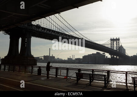 Man fishing from the East River Esplanade in New York City as the sun rises over the Manhattan Bridge Stock Photo