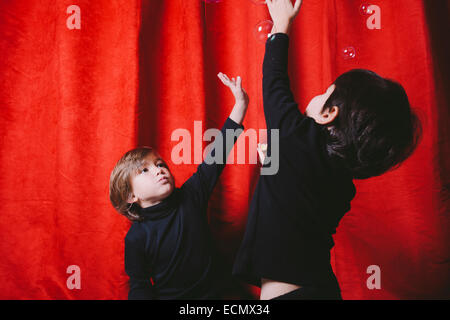 Two boys wearing black clothes playing with soup bubbles against a red curtain Stock Photo