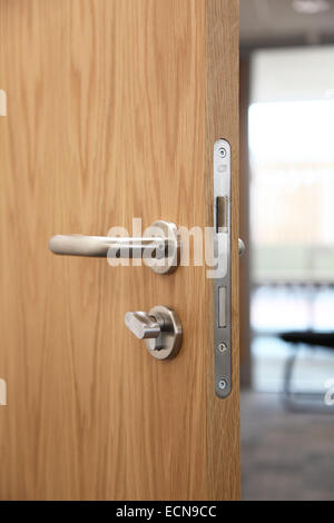 A partially open, modern, oak-finish interior door showing door handle, lock and mechanism and commercial interior beyond Stock Photo
