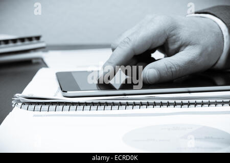 A tablet PC on an office table with an human hand pointing at it. There are spreadsheets on white computer paper covering desk Stock Photo