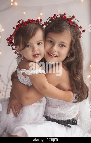 Girl hugs her little sister, both wearing red Christmas wreaths and smile Stock Photo
