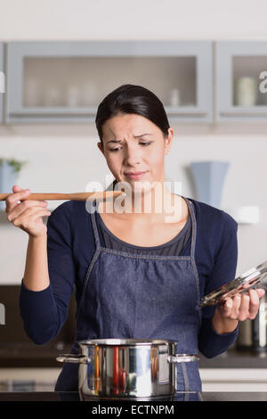 Attractive young female cook standing at the hob in her apron tasting her food in the saucepan with a grimace as she finds it di Stock Photo