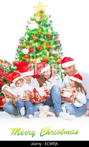 Happy family in Christmas eve at home sitting near decorated fir tree over white background, wearing red Santa hats Stock Photo
