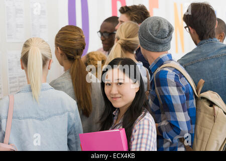 Portrait of smiling university student standing in corridor, people in background looking at exam results Stock Photo
