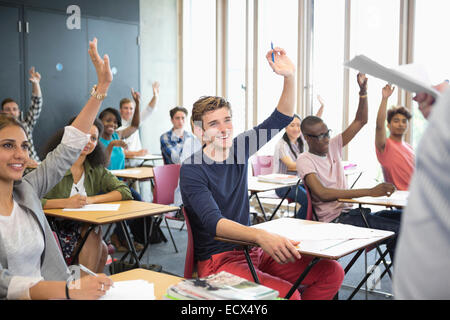 View of smiling students sitting at desks in classroom with arms raised Stock Photo