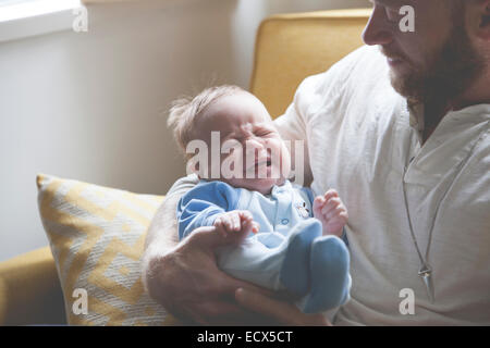 Father holding and looking at crying baby, sitting on sofa Stock Photo