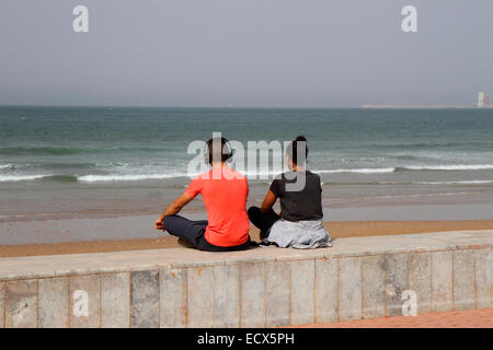 Young man and woman sitting on wall looking out to sea and wearing head phones Stock Photo