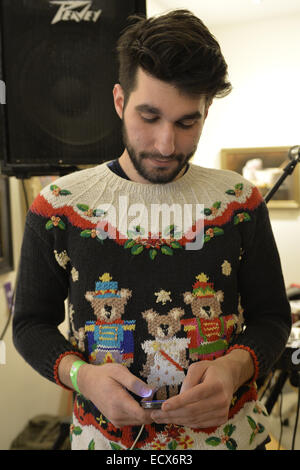 Huntington, New York, USA. 20th Dec, 2014. A young man wearing a festive, funny Christmas sweater decorated with three bears dressed for a parade looks down at his mobile phone, at the Jingle Boom Holiday Bash, which has entertainment, and prizes for people wearing the most creative or Ugly Sweaters, at the Main Street Gallery of Huntington Arts Council. Sparkboom, an HAC project, provides events such as this geared to Gen-Y, 18-34 years of age, to address the 'brain drain' of creative young professionals of Long Island. The paintings on the art gallery walls were the Annual Juried Stock Photo