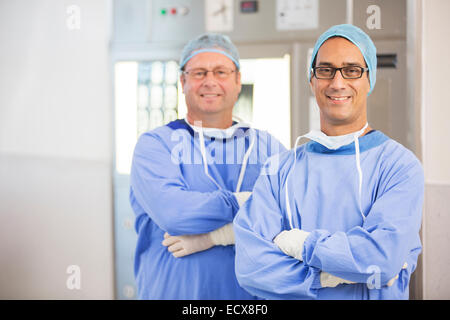Two smiling doctors with arms crossed, wearing surgical clothing and eyeglasses in hospital Stock Photo