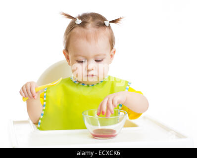 Baby eating and put fingers into food sitting at table Stock Photo