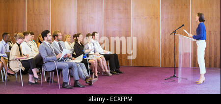 Young woman standing at transparent lectern, talking before audience in conference room Stock Photo