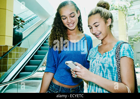 Mixed race teenage girls using cell phone near escalator at shopping mall Stock Photo