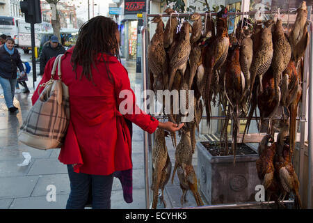 Woman checking the qulaity of pheasants hanging up outside a traditional British butchers shop on Bethnal Green Road, London, UK Stock Photo