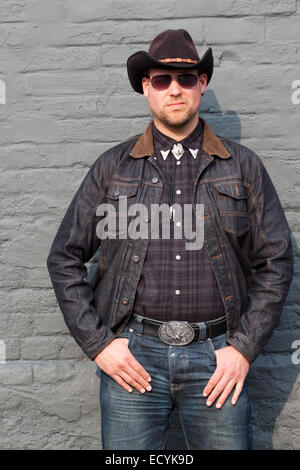 A young man dressed as a cowboy and wearing sunglasses poses in a macho way by a brick wall Stock Photo