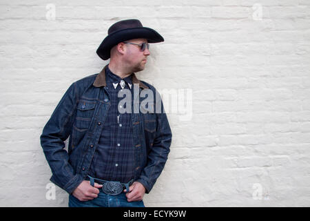 A young man dressed as a cowboy and wearing sunglasses poses in a macho way by a brick wall Stock Photo