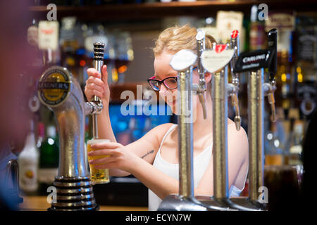 Part time job: A young person woman girl wearing spectacles working behind the bar in a pub public house pouring a pint of lager beer, UK Stock Photo