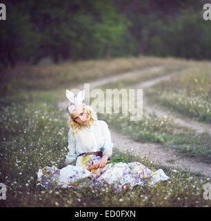 Young woman sitting in meadow dressed as a bunny holding a carrot Stock Photo