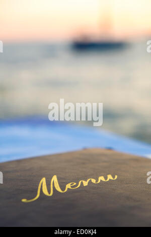 A menu laid on a seafood restaurant table with a boat out of focus on the sea at sunset in the background Stock Photo